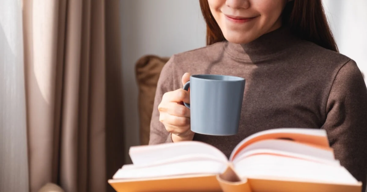 a woman smiling reading a book with a hot drink