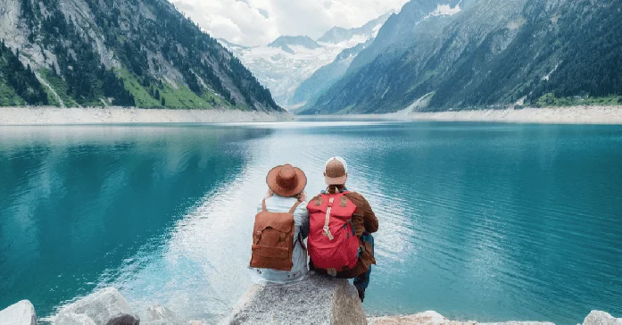 A travelling couple sit on a rock facing a clear blue lake in the mountains