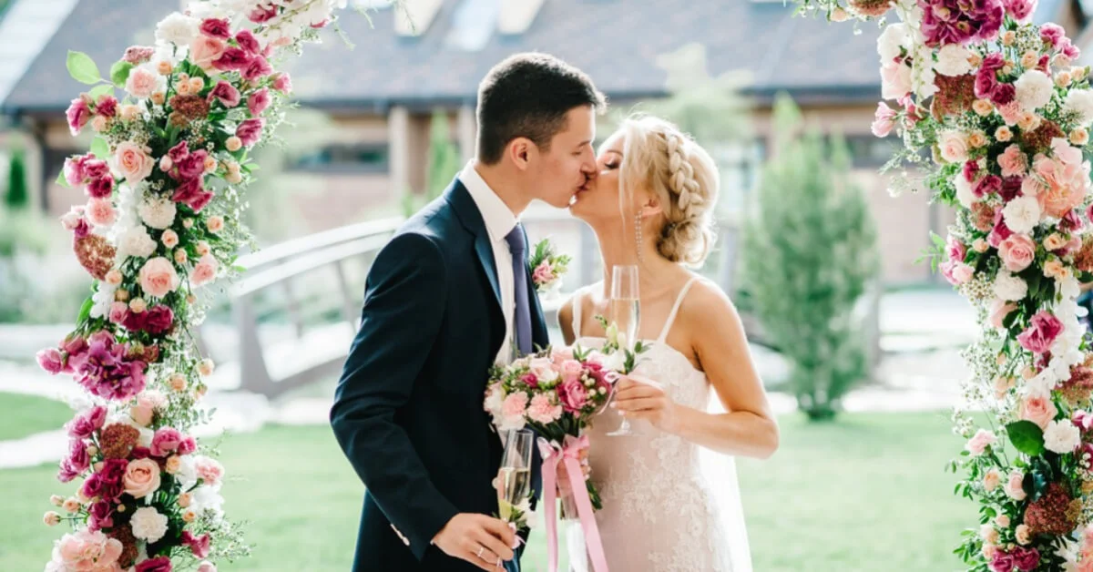 newly weds kissing under a flower arch holding a bouquet of flowers