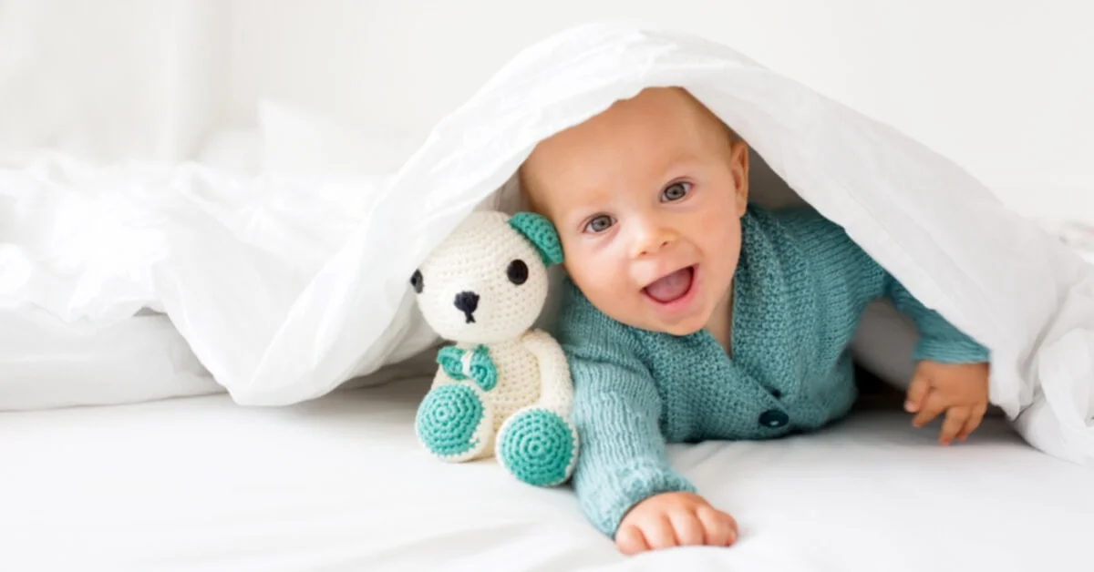 a baby smiling under a blanket with a knitted toy
