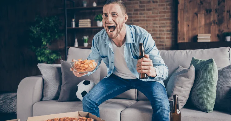Man shouting at the TV holding a beer and some crisps