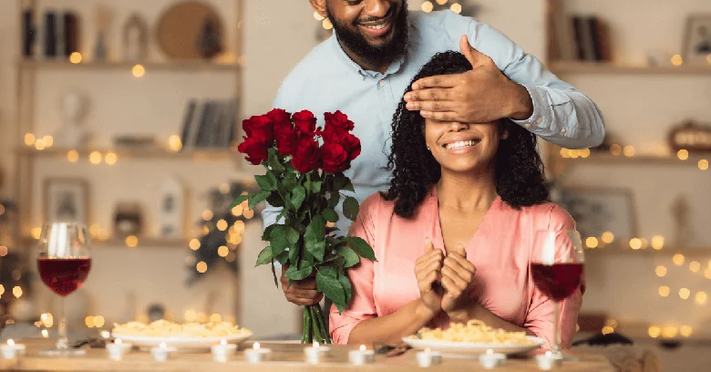 A man covering his wife or partners eyes at a table holding red roses